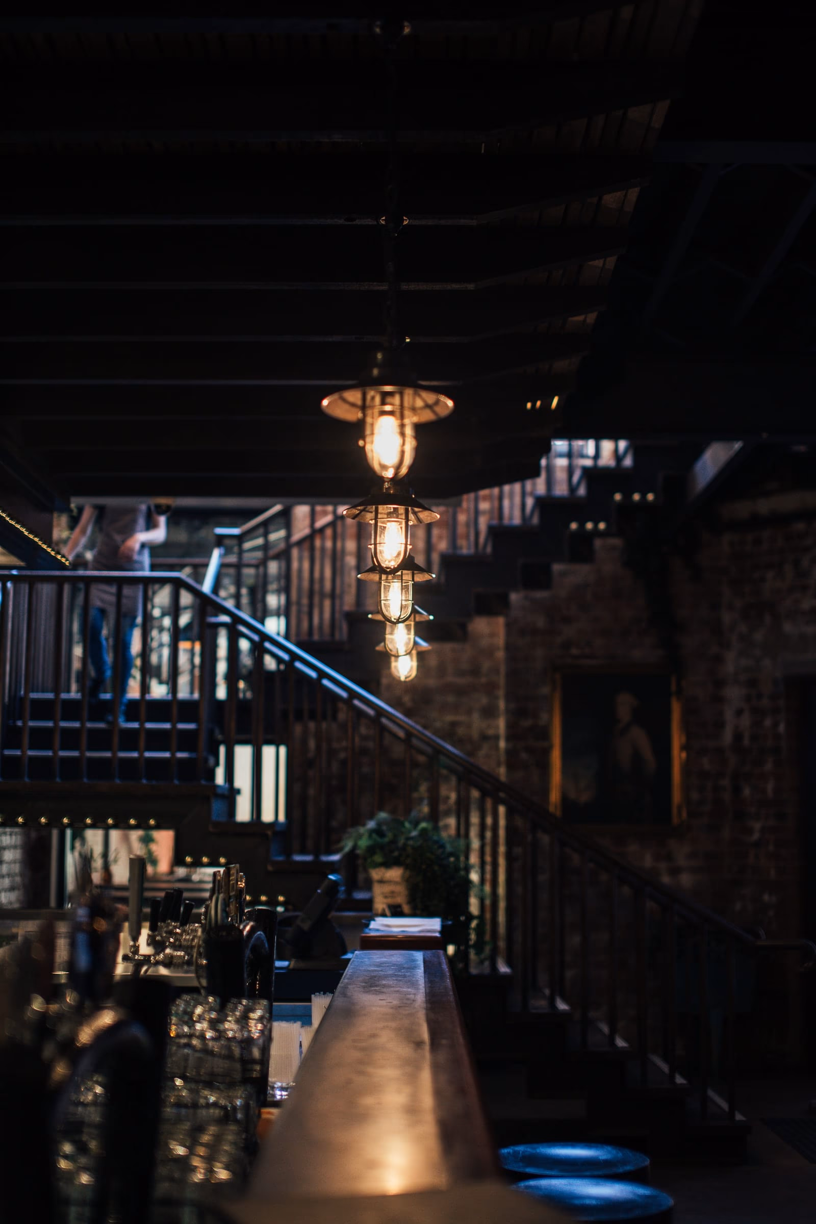 Caged pendants over a bar counter, a blackened-steel stair and exposed brick beyond