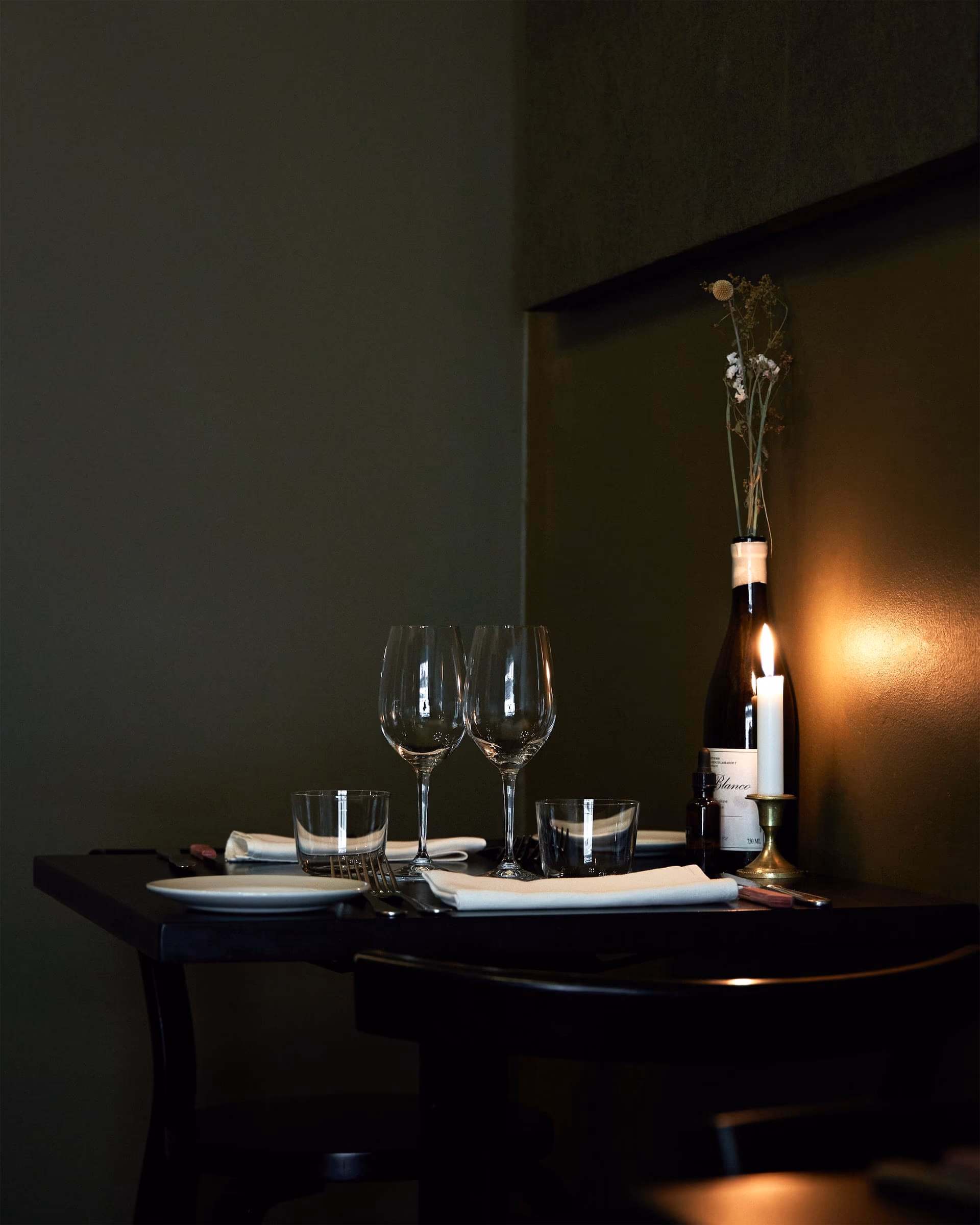 A table laid for two in near darkness, lit by a single candle against a deep olive wall